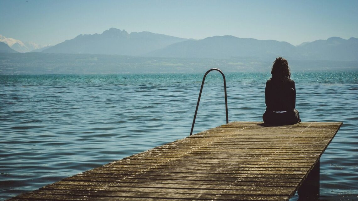 A solitary figure standing at the edge of a still lake at dawn, facing the water in quiet reflection