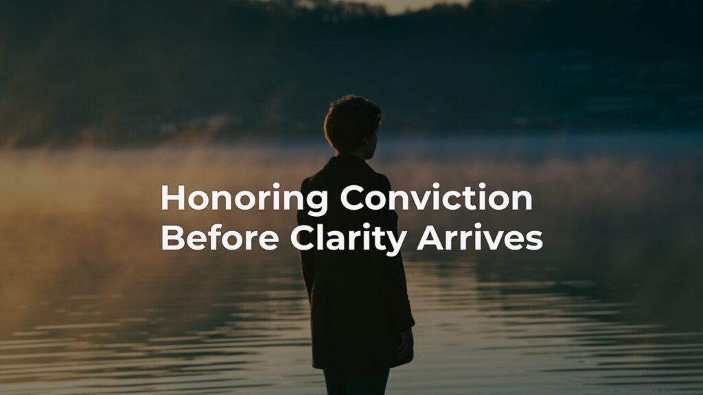 A person standing at the edge of a misty lake at dawn, honoring conviction before clarity by pausing to sense what lies ahead.