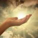 An open hand reaching gently toward sunlight through clouds