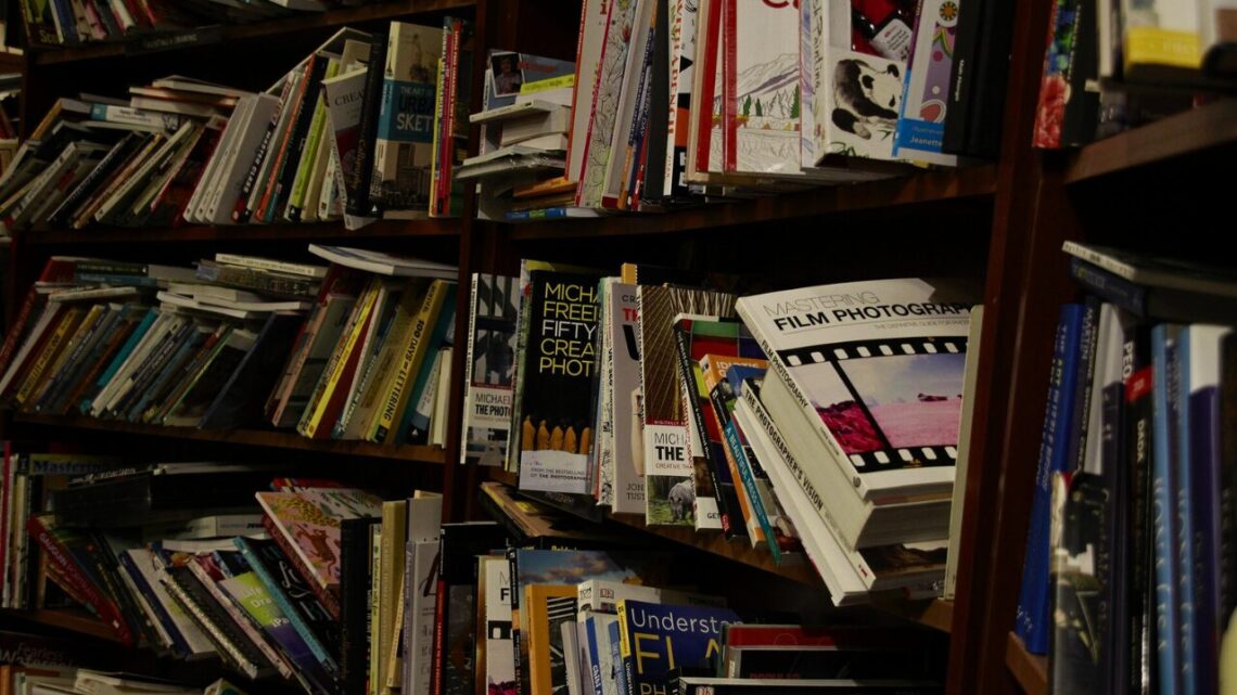 A densely packed bookshelf overflowing with books stacked and wedged in every direction, warm dim light casting deep shadows.