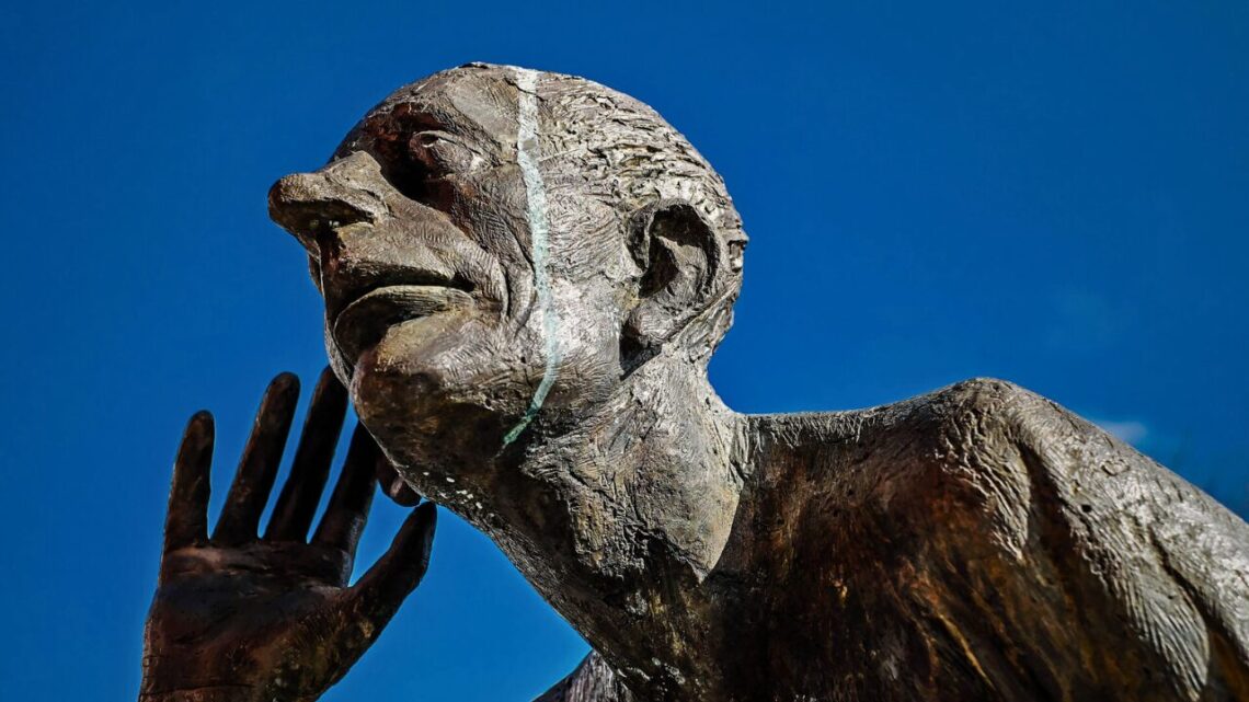 A weathered bronze statue of a man with hand to ear, gazing upward against a vivid blue sky, symbolizing deep attentive listening.