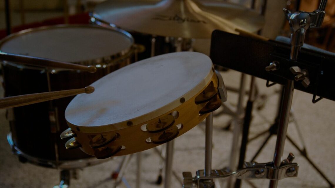 A tambourine and snare drum with drumsticks arranged in a dimly lit percussion setup, shot in close detail with shallow depth of field.