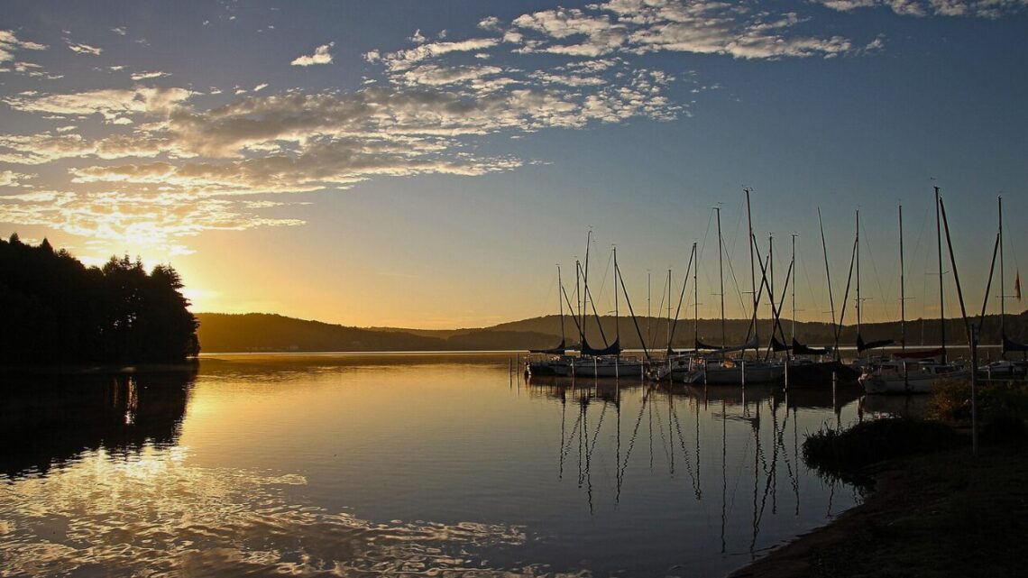 Sailboats moored at a calm marina at sunset, their masts reflected in still golden water beneath a wide sky with scattered clouds