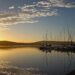 Sailboats moored at a calm marina at sunset, their masts reflected in still golden water beneath a wide sky with scattered clouds