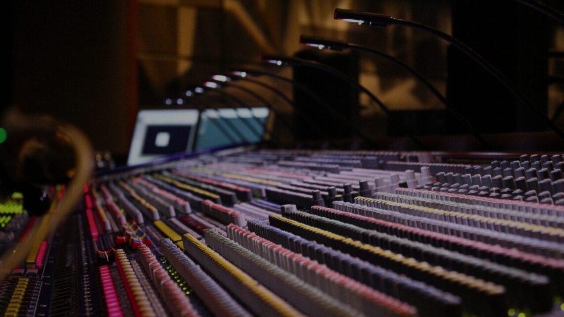 A large-format mixing console in a professional recording studio, rows of faders and channel strips stretching into soft focus, microphone booms and a monitor visible in the background