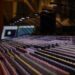 A large-format mixing console in a professional recording studio, rows of faders and channel strips stretching into soft focus, microphone booms and a monitor visible in the background