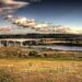 A wide road bridge spanning a tidal estuary, surrounded by golden fields and rolling green hills under a dramatic cloudy sky — the bridge Stephen crossed by bicycle at age twelve on his way to school.