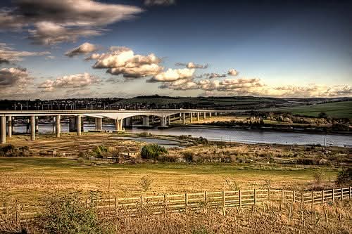 A wide road bridge spanning a tidal estuary, surrounded by golden fields and rolling green hills under a dramatic cloudy sky — the bridge Stephen crossed by bicycle at age twelve on his way to school.