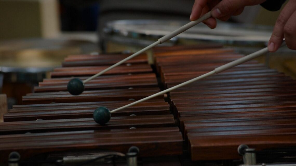 A musician's hands poised over a marimba with mallets mid-stroke, captured in a moment of focused, intentional presence during rehearsal.