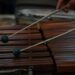 A musician's hands poised over a marimba with mallets mid-stroke, captured in a moment of focused, intentional presence during rehearsal.