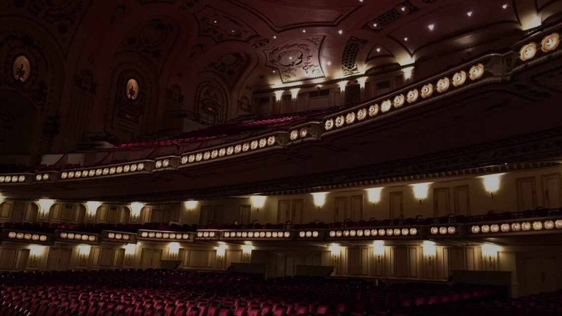 The ornate interior of an empty concert hall glows with warm stage lighting across tiered balconies and decorative plasterwork, the red seats waiting in stillness.