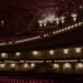 The ornate interior of an empty concert hall glows with warm stage lighting across tiered balconies and decorative plasterwork, the red seats waiting in stillness.