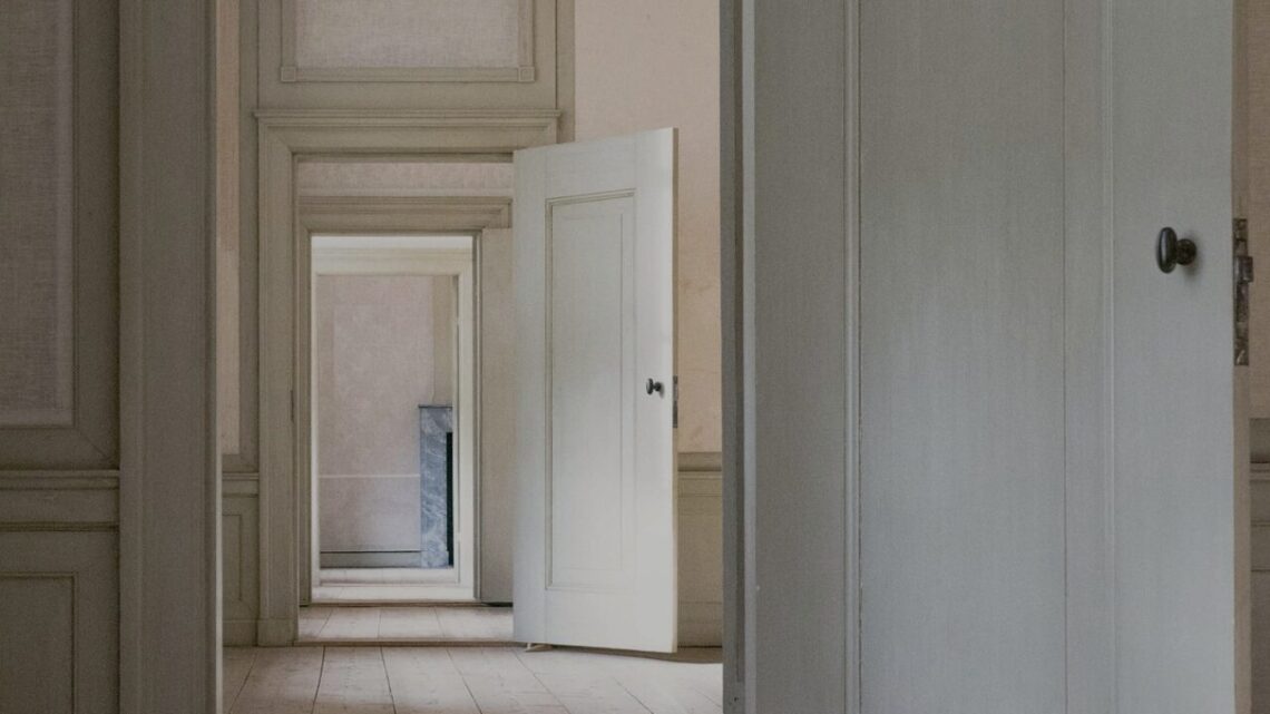 A receding sequence of open doorways through interconnected rooms in a classical interior, pale walls and worn wooden floors leading toward distant light, conveying purposeful forward movement through ordinary spaces.