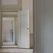 A receding sequence of open doorways through interconnected rooms in a classical interior, pale walls and worn wooden floors leading toward distant light, conveying purposeful forward movement through ordinary spaces.