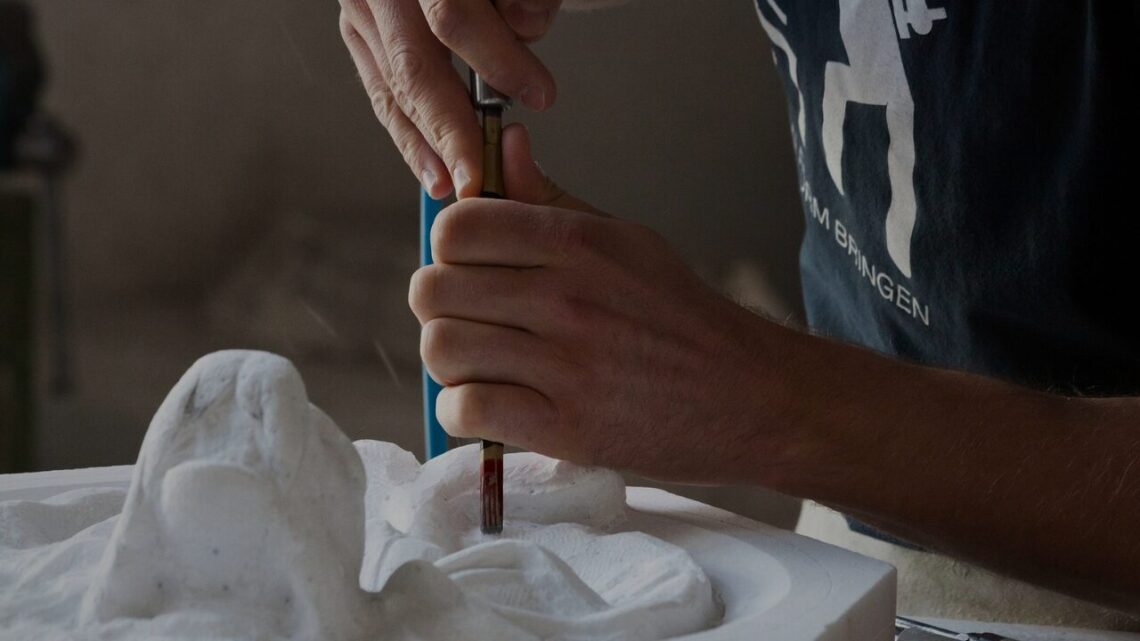 A sculptor's hands pressing a chisel into white stone, dust rising from the surface, partial figure visible in a dim workshop — the discipline of working within the properties of a medium to bring form into being.