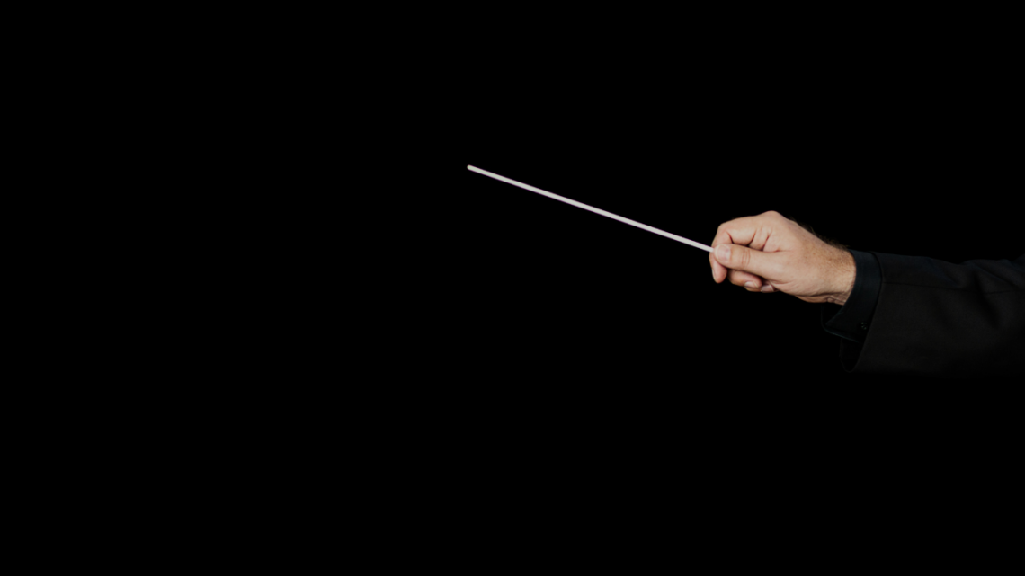 A conductor's hand holding a baton against a dark background, poised in stillness before the downbeat.