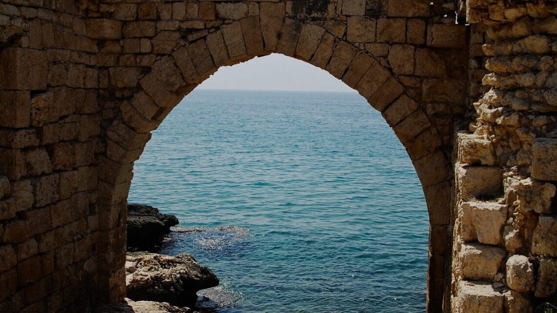 An ancient stone archway frames an open expanse of blue sea and distant coastline, warm sunlight catching the worn limestone edges.
