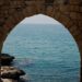 An ancient stone archway frames an open expanse of blue sea and distant coastline, warm sunlight catching the worn limestone edges.