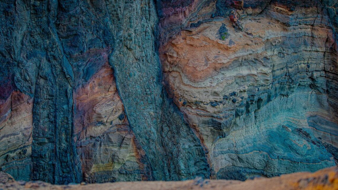 Dramatic layers of sedimentary rock in deep blue, terracotta, and cream reveal the visible record of accumulated time — each stratum a pattern deposited quietly over years.
