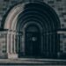 The Romanesque stone archway of a historic building, its layered concentric arch framing a darkened entrance, rendered in deep cool tones — a threshold held open, waiting.