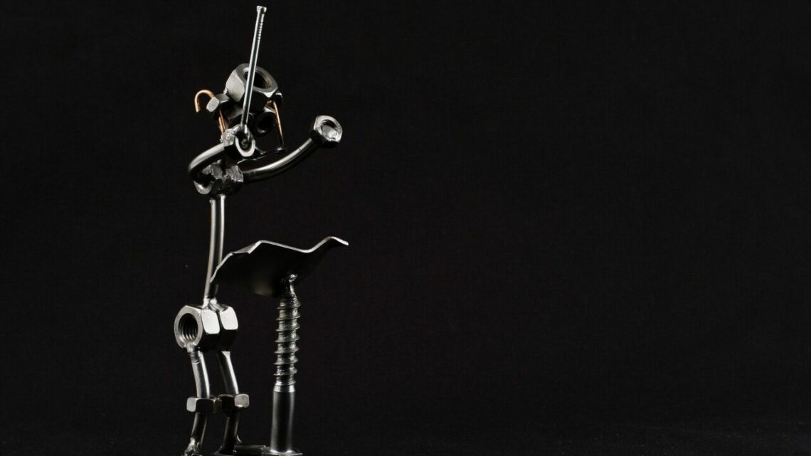 A miniature handcrafted sculpture of a conductor, made from nuts, bolts, and scrap metal, baton raised mid-gesture against a plain black background