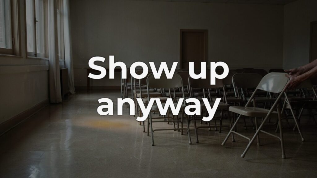 A pair of hands arranging folding chairs in rows inside a dim, empty fellowship hall lit by soft window light — the quiet courage of showing up before anyone else arrives.