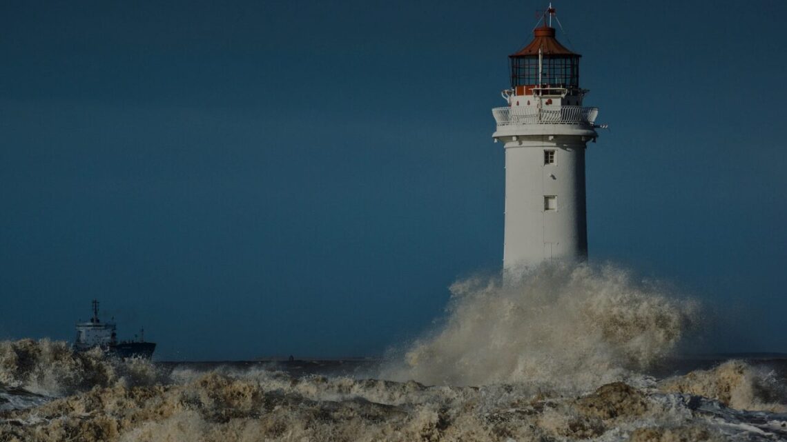 A white lighthouse with a red lantern room holds firm against a large breaking wave and a dark stormy sea, a cargo ship visible in the distance.
