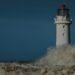 A white lighthouse with a red lantern room holds firm against a large breaking wave and a dark stormy sea, a cargo ship visible in the distance.