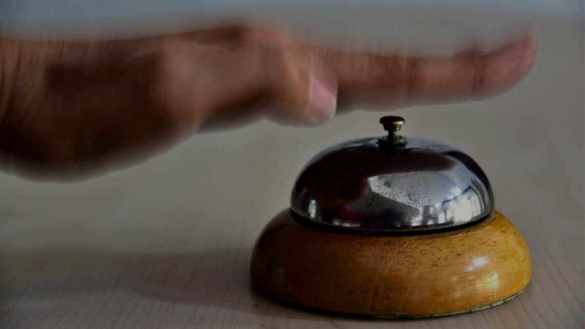 A hand reaches down to ring a worn brass service bell resting on a hotel reception desk.