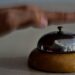 A hand reaches down to ring a worn brass service bell resting on a hotel reception desk.