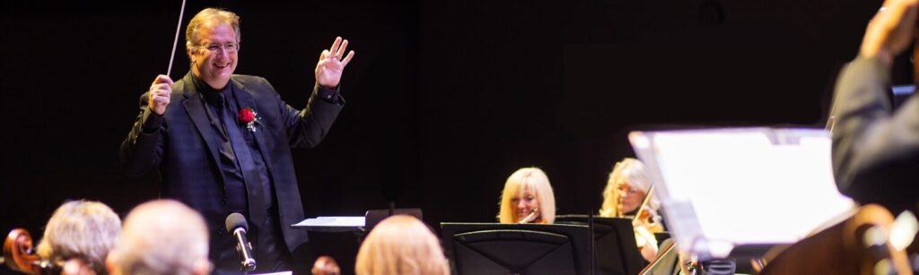 Stephen P. Brown conducting the Richey Community Orchestra, baton raised mid-cue, with the string section smiling in the foreground.