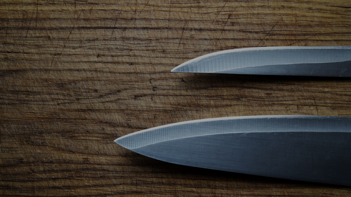 Two kitchen knives rest on a deeply worn wooden cutting board, its surface etched with years of the same daily strokes.