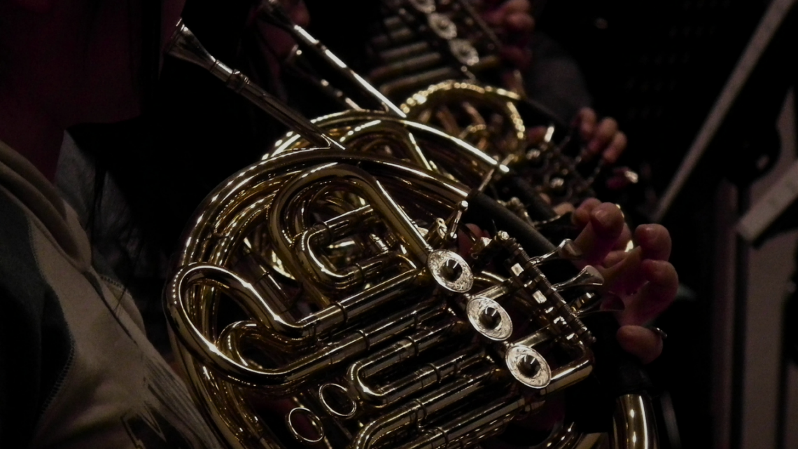 Close-up of French horns catching warm stage light in an orchestra section, valves and bell curves gleaming as a musician's hand rests on the keys.