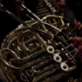 Close-up of French horns catching warm stage light in an orchestra section, valves and bell curves gleaming as a musician's hand rests on the keys.