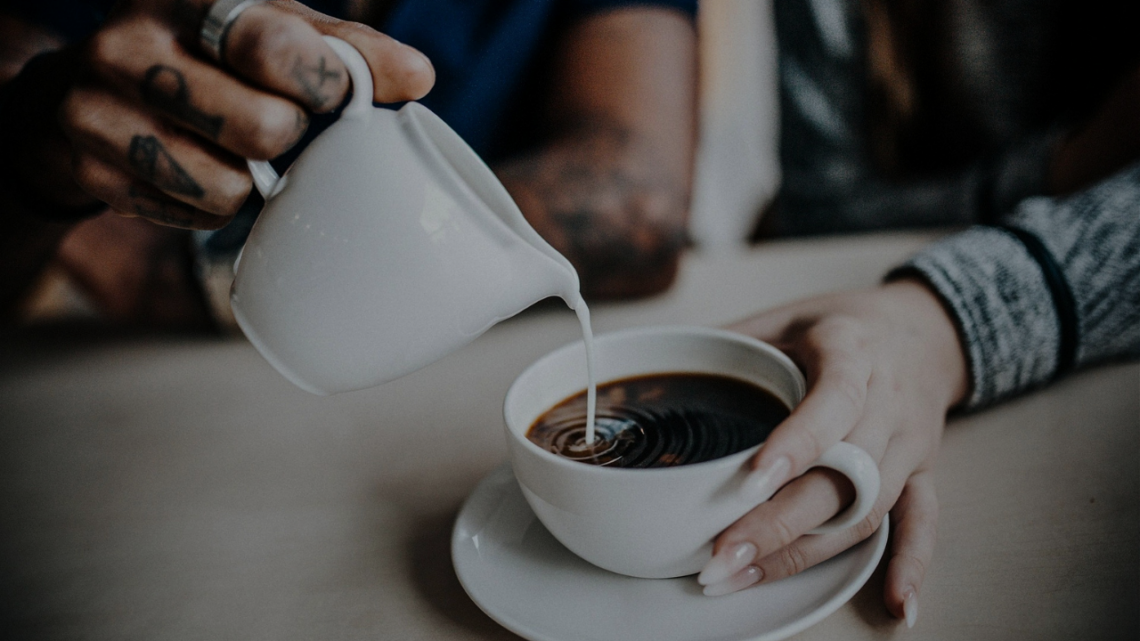 Hands pouring cream into a fresh cup of coffee, a small gesture of care handed to someone at the start of their day.