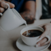 Hands pouring cream into a fresh cup of coffee, a small gesture of care handed to someone at the start of their day.