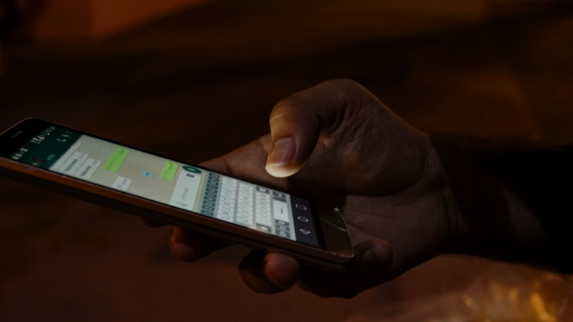 A hand holding a phone with a messaging app open, thumb hovering over the keyboard in dim light, a late-night message about to be sent.