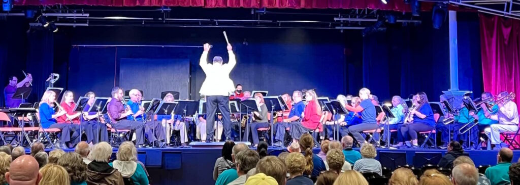 Stephen P. Brown conducting the Dunedin Concert Band, arms raised, with the audience and full ensemble visible.